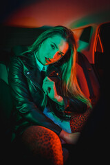 Urban photography with green and red neons in a car. Young blonde Caucasian model pose in a black leather jacket