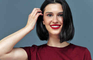 Fototapeta premium Horiozntal studio candid portrait of a young woman with red lips smiling broadly with white teeth. Brunette female model with red lips has joyful expression on blue background.