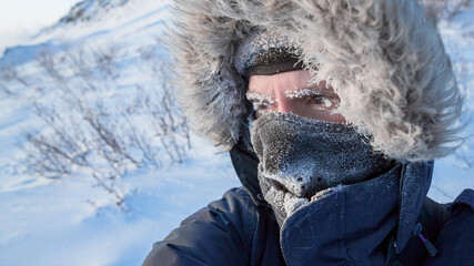 Portrait of a traveler in the Arctic. A man in winter clothes, a hood and a mask. Ice on the eyebrows and eyelashes. Extremely cold weather in the far north in the Arctic. Travel to the polar region. © Andrei Stepanov