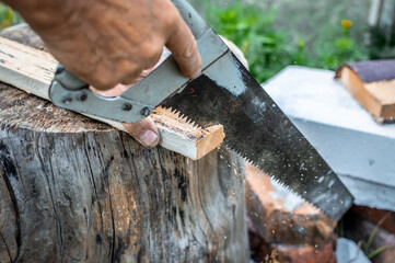 A man's hand uses a hand saw or hacksaw to cut a piece of a wooden board leaning on a stump. Outdoor carpentry