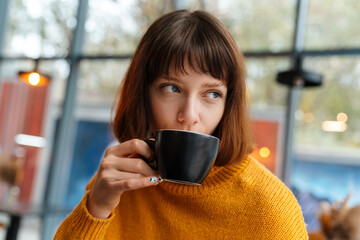 Beautiful redhead girl drinking coffee while sitting in cafe indoors