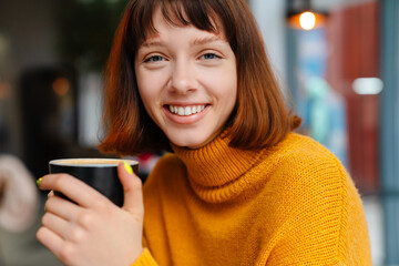 Happy redhead girl smiling and drinking coffee while sitting in cafe