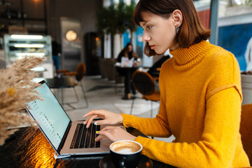 Focused redhead girl drinking coffee and working with laptop in cafe