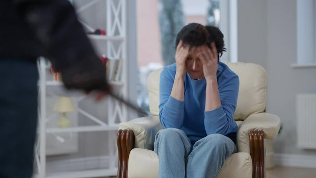 Desperate Caucasian woman holding head in hands asking murderer not to kill her. Portrait of stressed hopeless victim talking to maniac sitting on armchair at home. Murder and crime concept.