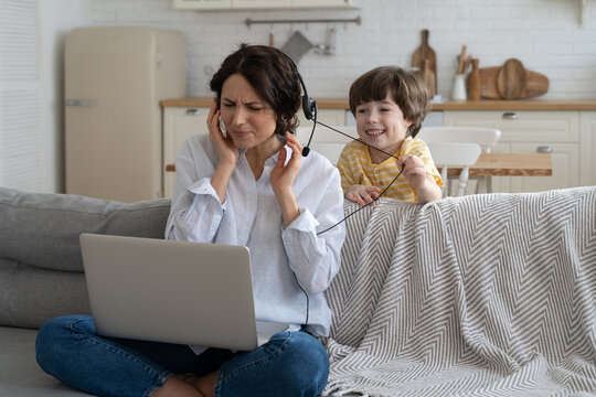 Nervous Freelancer Mother Sitting On Couch At Home Office During Lockdown, Working On Laptop. Little Child Distracts From Work, Taking Off Headphones, Making Noise And Asking Attention From Busy Mom