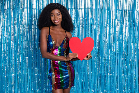 Portrait of attractive cheerful girl holding in hands paper heart 14 February romance isolated over blue tinsel curtain background