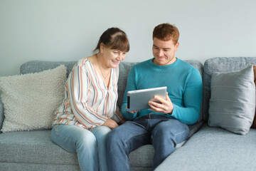 son teaching his mother to use tablet. older people using technology. Cheerful elderly woman sitting on the sofa next to his adult son