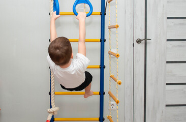 An active little boy does sports exercises on stairs of house. A small preschool boy does morning exercises on home horizontal bar