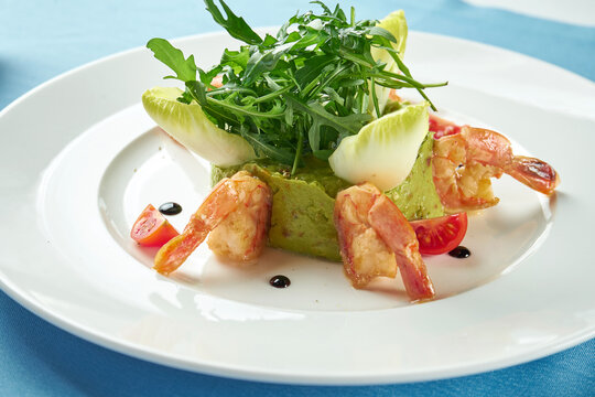 Avocado guacamole with shrimp, arugula and cherry tomatoes in a white plate on a blue tablecloth. Close up. Mexican food