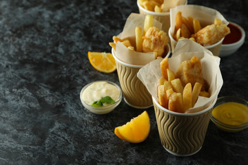Paper cups with fried fish and chips, and sauces on black smokey background