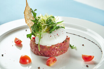 Delicious appetizer - beef tartare with croutons, poached egg and arugula salad in a white plate on a blue tablecloth