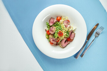 Appetizing, warm salad with roast beef, cherry tomatoes, parmesan and pine nuts in a white plate on a blue tablecloth