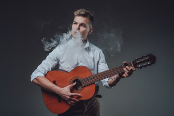 Obraz premium Portrait of a hipster guy dressed in formal clothing playing guitar in dark background and smoking cigarette.