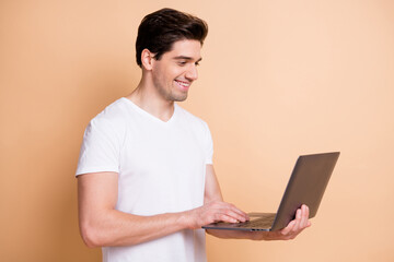 Portrait of half turned handsome cheerful guy using netbook write message isolated on beige color background