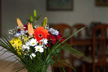 Bouquet of flowers in a glass vase in a living room as a decoration with chairs, tables and pictures in the background. colorful flowers