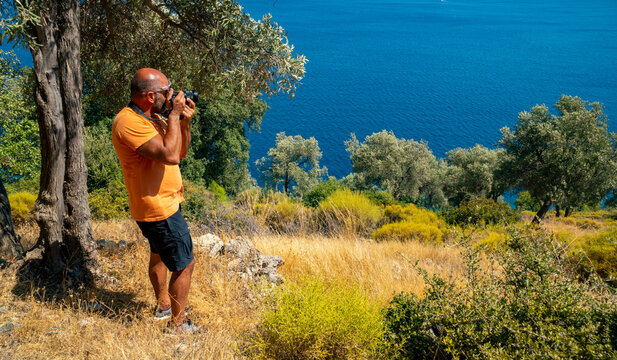A casual Caucasian male photographer is taking pictures in nature. He is holding a black big camera. The adult male is outdoors alone and free on a summer day 