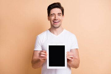 Portrait of funny friendly man arms hold tablet showing display eye wink isolated on beige color...