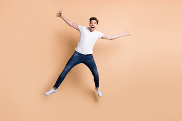 Fototapeta na wymiar Full size photo of optimistic brunet man jump yell wear t-shirt jeans sneakers isolated on beige color background