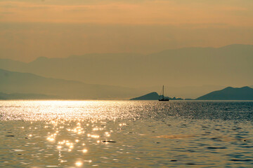A single sailing boat floating on tranquil waters of the Aegean Sea during sunrise or sunset. Beautiful sun is shimmering golden on waves. Islands in background with copy space