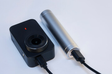 A compact action camera for shooting video and photos on a white background is charged from a power bank.