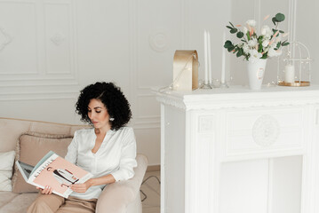 A woman with curly hair sits on a sofa and reads a Kinfolk magazine next to the fireplace