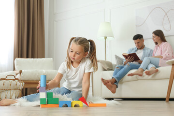 Little girl playing with colorful blocks while her parents resting on sofa at home. Floor heating...