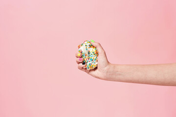 Unhealthy food. Female hand squeezing donut with colored sprinkles on pink background