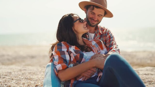 happy romantic couple sits on sea beach, talks, laughs and hugs. handsome adult man in casual checkered shirt and hat embracing beautiful charming young woman. moments of love, tender and happiness