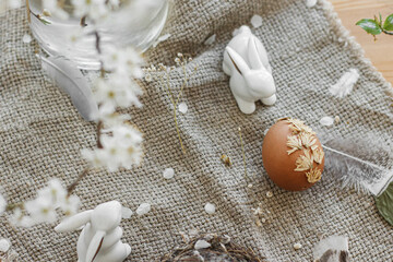 Easter egg decorated with dry flower and bunny on rustic table with linen napkin, feather and bloom