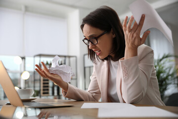 Emotional businesswoman with crumpled paper working on laptop in office. Online hate concept