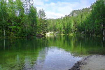 Fototapeta premium The green forest is reflected in the water. Rock on the lake shore. Summer landscape