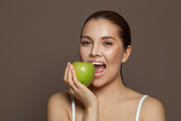 Attractive young woman biting green apple and smiling on brown background