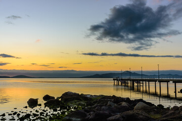 Seascape at golden hour on the horizon, pier and rocks in the foreground, golden sky and clouds - 419612211