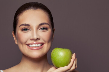 Beautiful woman with cute smile with white teeth smiling and holding green apple on brown background