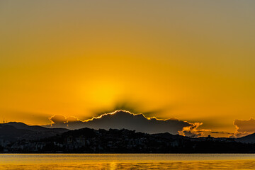 Sunrise behind clouds with golden sky, hills on the horizon and golden sea - 419613014