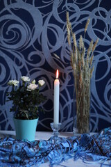 Burning white candle on blue textile with roses in pot and dry wheat spikes in vase