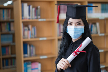 A young happy cheerful Asian woman university graduates in graduation gown and cap wears a face mask holds and shows a degree certificate to celebrate her education achievement on the commencement day