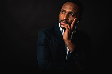 Portrait of attractive, handsome, stylish professional African American businessman in stylish suit and white shirt pensive holding hand near face, isolated on dark background. Low key. Selective focu