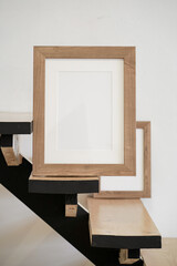 Closeup of square empty wooden picture frame on wooden stairs. White wall background. Scandinavian interior, home design. Art concept. Artistic mockup scene. Front view