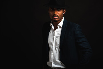 Portrait of attractive, cute, stylish professional African American businessman in stylish suit, white shirt and black hat, isolated on dark background. Low key. Selective focus