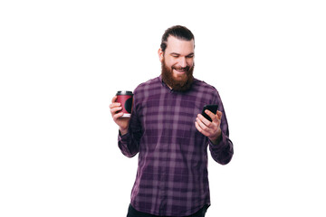 Cheerful young happy bearded man holding cup of coffee to go and using smartphone