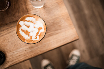 A hot cup of latte art coffee on a wooden table where people stand wearing blue sneakers on the floor at the coffee shop