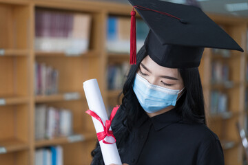A young happy cheerful Asian woman university graduates in graduation gown and cap wears a face mask holds and shows a degree certificate to celebrate her education achievement on the commencement day
