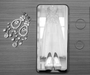 wedding accessories, a phone with a photo of a wedding dress next to earrings and wedding rings. Black and white photo.
