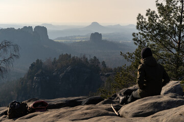 A man sitting on a huge rock at the edge of a cliff on a mountain top and looking down to the slightly hazy valley with impressive rock formations in the Sächsische Schweiz, Dresden