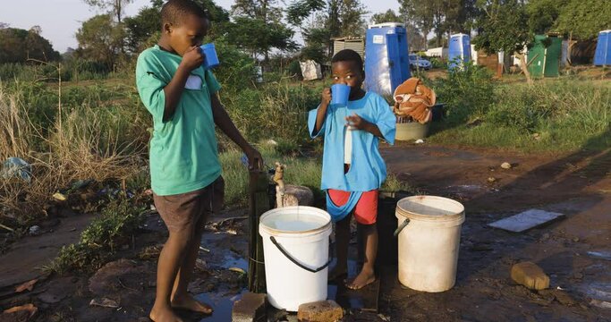 Water crisis. Close-up view of two young black African boys drinking water from a communal faucet and filing up plastic containers to take back home due to poor living conditions, no running water  