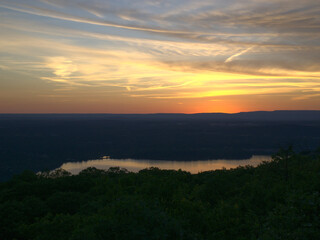 Thick forest, countryside town, orange and yellow sunset reflecting on a lake and cloudy sky with multiple illuminated contrails, Breakneck Ridge Trail, Hudson Highlands, New York, United States