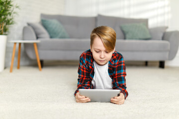 child boy in checkered shirt lies on floor and looks at the tablet. kid using digital tablet at home