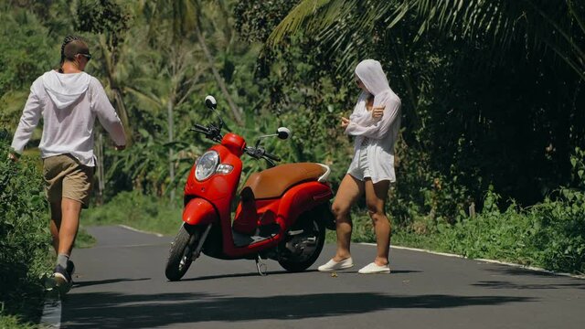 Two caucasian tourist woman man dance near scooter. Love couple on red motorbike in white clothes, sunglasses on forest road trail trip. Dancing road. Motorcycle rent. Asia Thailand ride tourism.