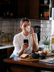 Beautiful woman drinking coffee in the morning. Smiling woman using the phone in the kitchen..